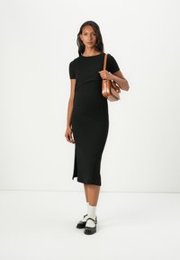 Μαύρο ριχτό midi φορεμα με κοντά μανίκια και πλάγιο άνοιγμα, συνδυασμένο με μαύρα παπούτσια, λευκές κάλτσες και μια καφέ τσάντα χιαστί.