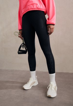 Black leggings with a smooth texture, featuring a patterned panel on the lower leg. Paired with white shoes and a bright pink sweatshirt.