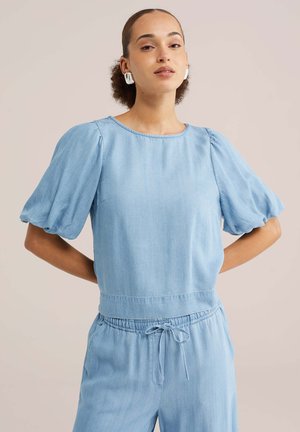 Žena s kučeravými vlasmi stiahnutými dozadu, oblečená v svetlomodrej blúzke s nafúknutými rukávmi a zodpovedajúcich nohaviciach so šnúrkou na stiahnutie na jednoduchom pozadí.