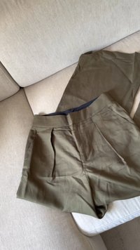Olivgrüne Hose aus einem strukturierten Stoff, mit hohem Bund, vorderen Falten und taillierter Passform, auf einem hellen Sofa ausgelegt.