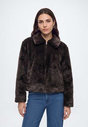 Jeune femme portant une veste en fausse fourrure marron foncé avec col et un jean bleu, debout devant un fond uni.