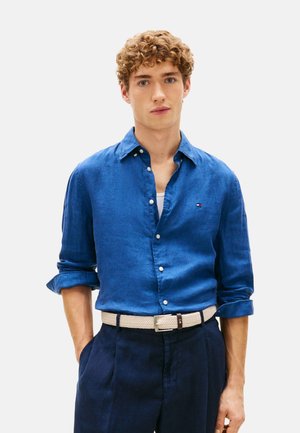 Joven con pelo rizado que lleva una camisa azul abotonada con las mangas remangadas, pantalones oscuros y un cinturón tejido claro, con las manos en los bolsillos.