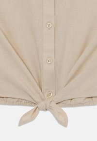 Beige knappskjorte med knyttet frontfalda. Laget av lett stoff, med fire kremfargede knapper og subtil tekstur.