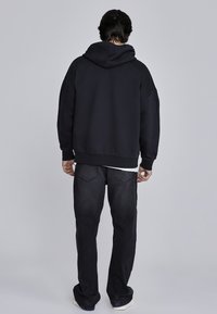 �Čierna mikina s veľkou kapucňou, spadnutými ramenami a rebrovanými manžetami, v kombinácii s tmavými džínsy a bielym tričkom, zobrazená zozadu.