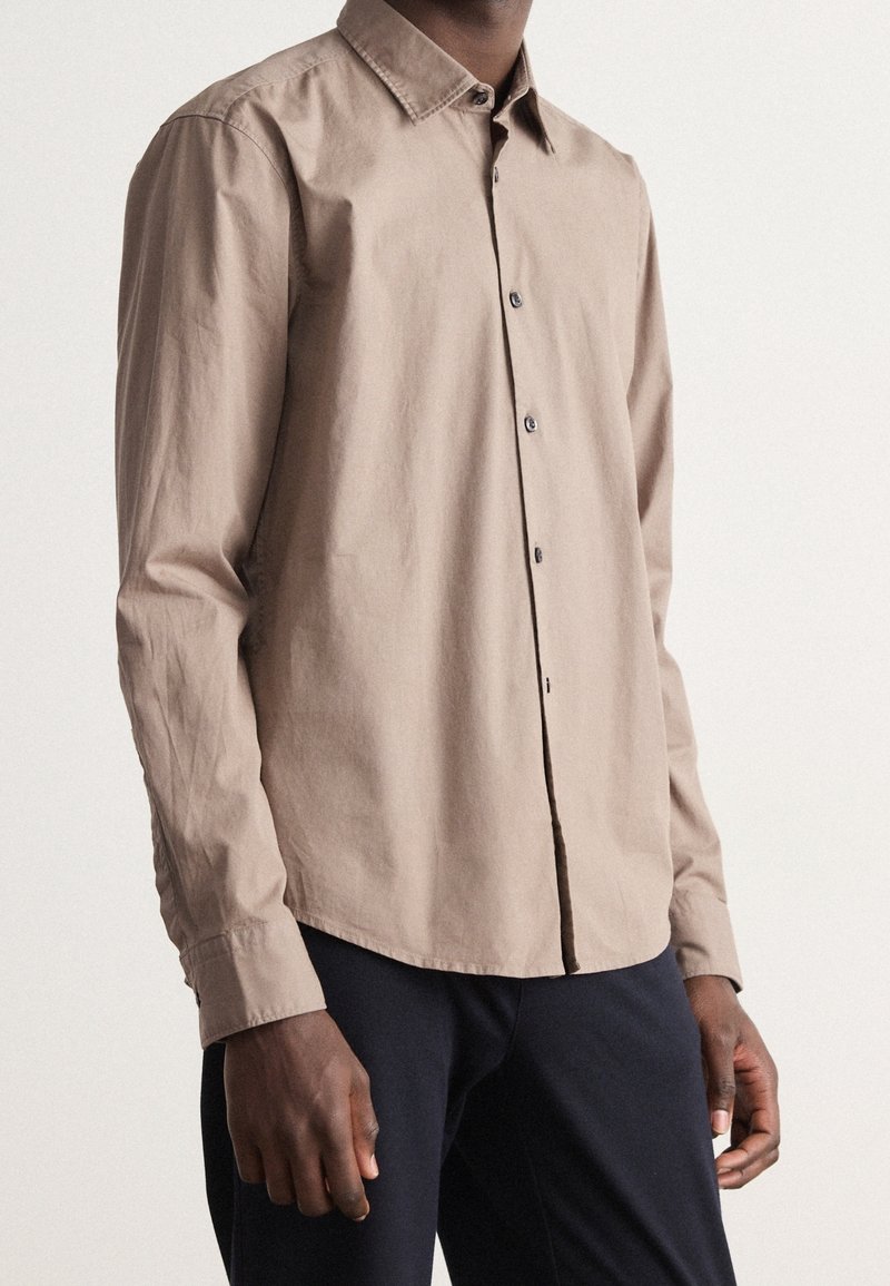 Hombre con camisa de botón de manga larga beige y pantalones oscuros, de pie frente a un fondo claro y liso.