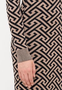 Main reposant contre un tissu tricoté géométrique beige et noir à motif avec un poignet côtelé sur la manche.