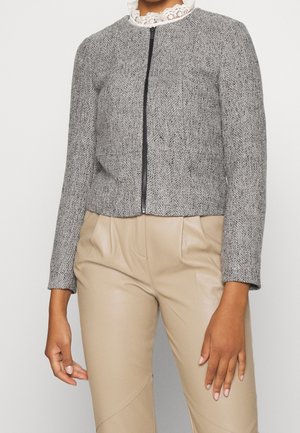 Femme portant une veste grise texturée zippée sur un top blanc à col en dentelle et un pantalon beige taille haute, debout devant un fond blanc.