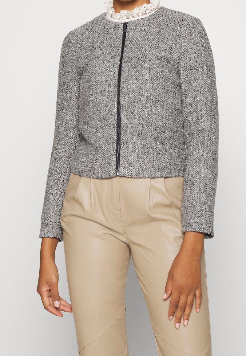Femme portant une veste grise texturée zippée sur un top blanc à col en dentelle et un pantalon beige taille haute, debout devant un fond blanc.