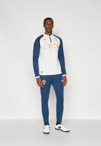 Λευκή και ναυτική αθλητική φόρμα με πορτοκαλί λογότυπο "INEOS", μακριά μανίκια, φερμουάρ στο ένα τέταρτο, στενά παντελόνια και λευκά αθλητικά παπούτσια.