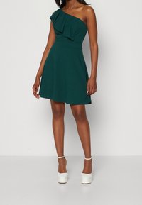 Grön, enaxlad klänning med volangutskärning, åtsittande midja och utsvängd kjol; ihop med vita klackskor med plattform. Slät tygtextur.