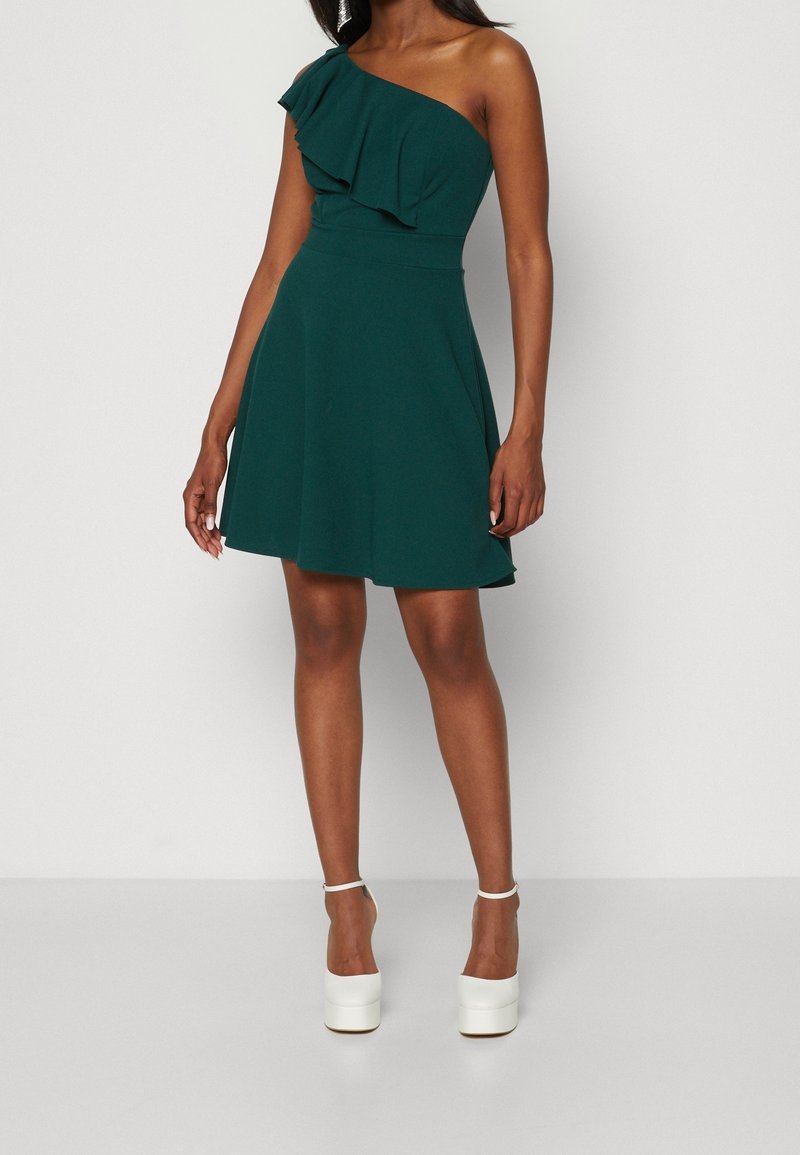 Grön, enaxlad klänning med volangutskärning, åtsittande midja och utsvängd kjol; ihop med vita klackskor med plattform. Slät tygtextur.