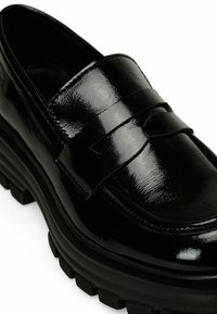 Mocassino in vernice nera con finitura lucida, caratterizzato da una punta arrotondata, doppi accenti a strappo e una suola in gomma nera spessa con grip.