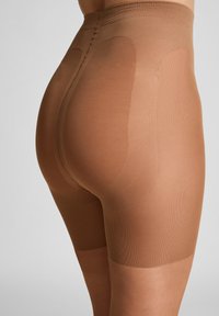 Nahaufnahme einer Person, die transparente, hautfarbene formende Shorts mit verstärkten Teilen an Hüften und Gesäß trägt, vor einem schlichten Hintergrund.