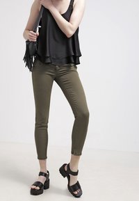 Haut noir sans manches avec ourlet à volants, pantalon ajusté vert olive, et sandales noires épaisses à larges brides. Sac bandoulière avec détail à franges.