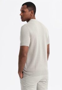 Vaaleanharmaa lyhythihainen polo-paita, jossa on teksturoitu neulekuvio ja ribbatut hihansuut, yhdistettynä vaaleisiin housuihin.