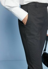 Dunkelgraue, maßgeschneiderte Hosen mit glatter Textur, ausgestattet mit Seitentaschen und einem Bund mit Gürtelschlaufen, kombiniert mit einem weißen, umgeschlagenen Hemd.