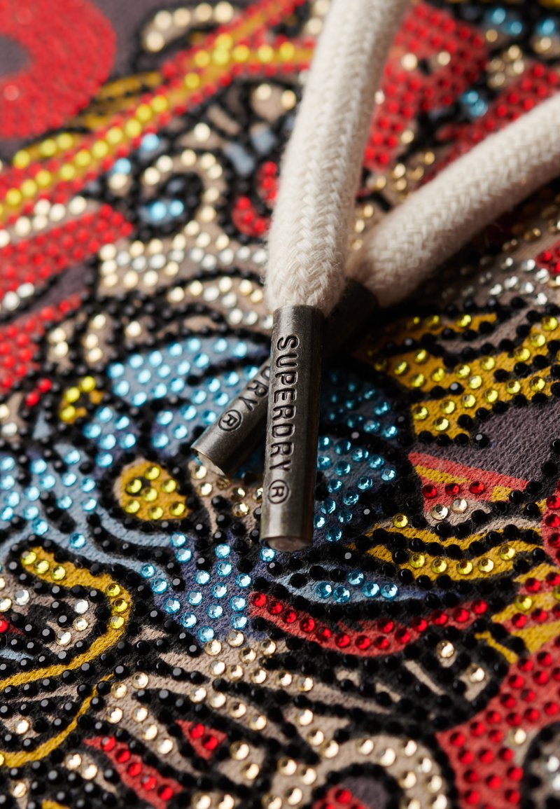 Zbliżenie beżowych sznurowadeł z metalowymi końcówkami z wytłoczonym napisem "SUPERDRY®" na kolorowej tkaninie ozdobionej czerwonymi, niebieskimi, żółtymi i czarnymi koralikami.
