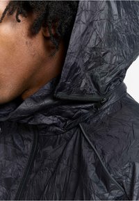 Schwarze, gewellte Stoff-Hoodie-Jacke mit Reißverschluss und verstellbarem Kragen. Hat minimale Hardware und eine strukturierte Oberfläche.