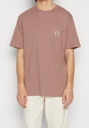 Ruskea puuvillainen t-paita, jossa on pyöreä kaula-aukko, lyhyet hihat ja rintatasku, jossa on neliönmuotoinen logopatchi. Yhdistetty vaaleisiin housuihin.