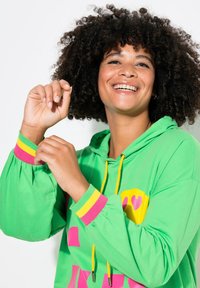 Felpa verde con strisce decorative rosa e gialle sulle maniche, con una stampa grafica. Realizzata in tessuto morbido, con una vestibilità comoda e cappuccio regolabile.