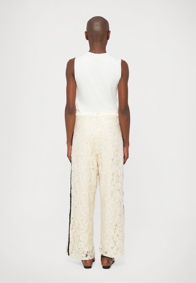 Top branco sem mangas combinado com calças largas de renda em creme com riscas pretas nas laterais. Textura suave e design fluido.