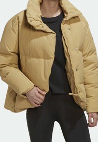 Personne portant une doudoune beige sur une chemise noire et un pantalon noir, main tenant la veste près de la fermeture éclair, vue de face recadrée au niveau du cou.