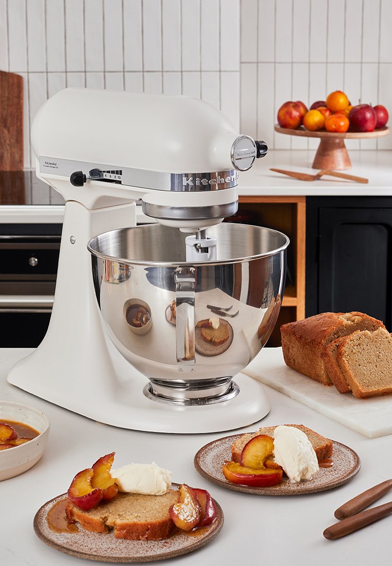 Weißer KitchenAid-Standmixer mit Edelstahlschüssel auf der Arbeitsplatte, umgeben von geschnittenem Kuchen, Pfirsichbelägen und Schälchen mit Soße in einer Kücheneinrichtung.