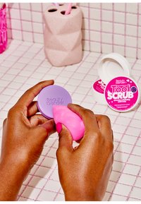 Gąbka Beauty Blender w różowym lateksie, trzymana blisko okrągłego fioletowego pojemnika oznaczonego "Beauty Blender". Tło ma różowy wzór w kratkę i dodatki.