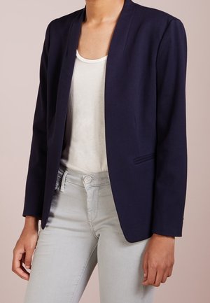 Femme portant un blazer bleu marine sur un t-shirt blanc à encolure dégagée et un pantalon gris clair, debout devant un fond beige uni.