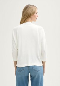 Witte gebreide trui met driekwart mouwen en een ronde halslijn, gecombineerd met lichtblauwe jeans, van achteraf getoond.