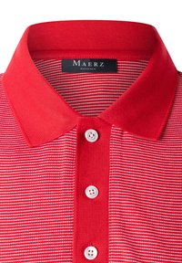 Röd polo-tröja med en texturerad krage, med horisontella vita ränder och tre knäppningar vid nacken. Varumärkesetikett synlig.