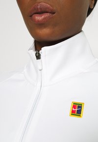 Hvid lynlåsjakke med høj krave, prydet med et flerfarvet broderet logo på venstre side, glat tekstur og atletisk design.