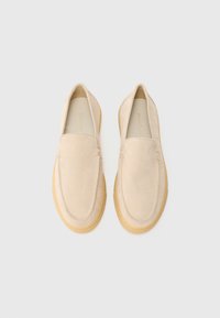 Par de mocasines de ante beige con puntas redondeadas y suelas de goma marrón, exhibidos lado a lado sobre un fondo blanco.