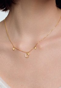 Złoty naszyjnik łańcuszkowy z dwoma zawieszkami w kształcie gwiazdek i zawieszką w kształcie półksiężyca z rzędem małych, przezroczystych kamieni, spoczywający na obojczyku.