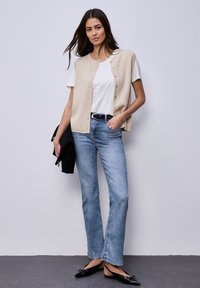 Femme debout contre un mur gris, portant un cardigan sans manches beige sur un t-shirt blanc, un jean bleu clair, une ceinture noire et des ballerines noires pointues, tenant une veste noire.