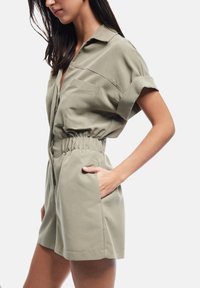 Tuta verde oliva con maniche corte, chiusura frontale con zip, vita elastica e due tasche laterali, realizzata in cotone leggero.