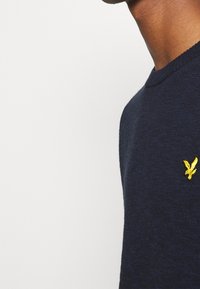 Närbild av en person som bär en mörkblå tröja med ett litet gult fågelmärke på bröstet, enkel vit bakgrund.