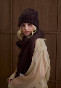 Bonnet tricoté marron foncé à texture côtelée, assorti à une écharpe épaisse de la même couleur. Le mannequin porte un haut léger beige transparent. Fond en bois.