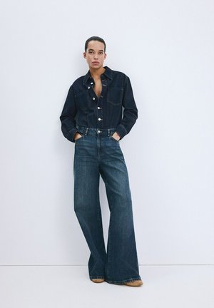 Persona con camisa de mezclilla oscura abotonada y vaqueros de pierna ancha, de pie con las manos en los bolsillos contra un fondo blanco liso.