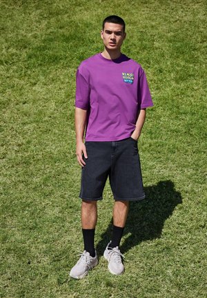 Nuori mies seisoo ruohikolla, pukeutuneena violettiin "Venice Beach Hip Hop" -paidassa, mustiin shortseihin, mustiin sukkiin ja valkoisiin lenkkareihin.