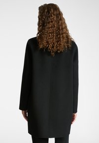 Cappotto di lana nero con maniche lunghe, taglio dritto e colletto alto, con una texture liscia e senza chiusure visibili.