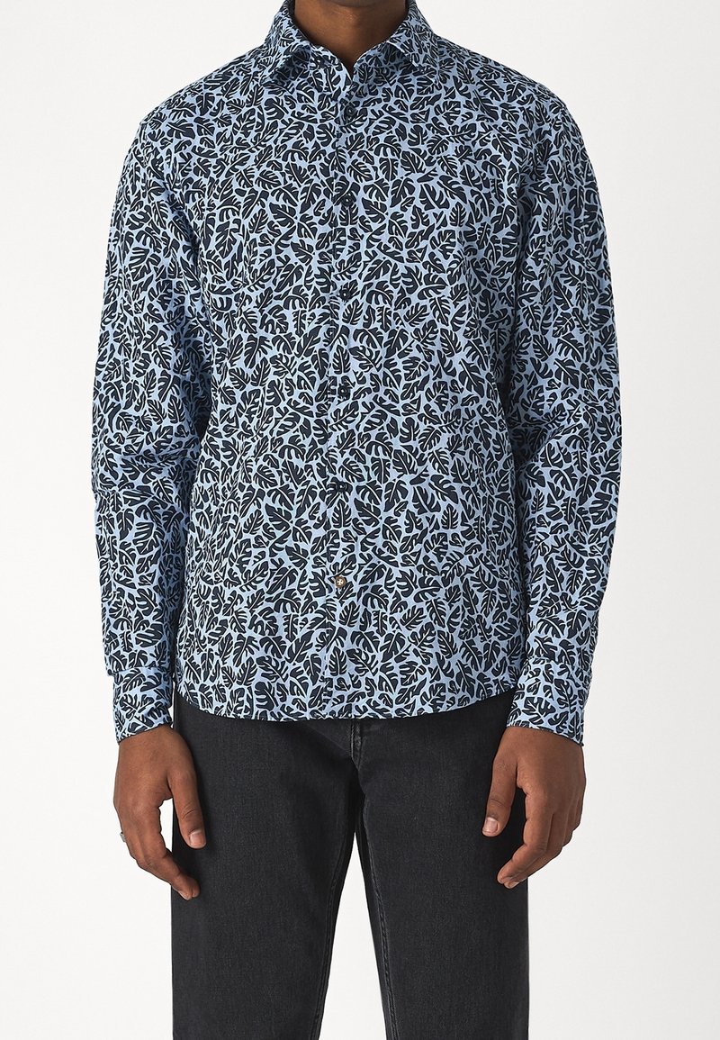 Homme portant une chemise boutonnée à manches longues avec un motif de feuilles bleu et noir, associée à un pantalon noir, debout sur un fond blanc.