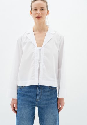 White long-sleeve blouse with a deep V-neck, tie closure, and a slightly cropped fit, paired with blue high-waisted jeans.