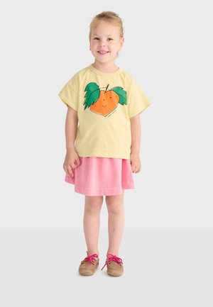 Jeune fille souriante debout, portant un t-shirt jaune avec un motif de fruit orange et de feuilles vertes, une jupe rose et des chaussures marron à lacets roses.