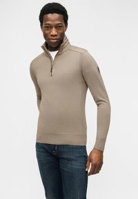 Άνδρας που φοράει μπεζ πουλόβερ με φερμουάρ στη μέση και σκουρόχρωμο μπλε τζιν, στέκεται μπροστά σε λευκό φόντο, κοιτάζοντας μπροστά με ελαφρύ χαμόγελο.