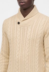 Homme portant un pull beige en maille torsadée avec un col châle et un seul bouton foncé près du cou.
