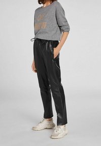Donna che indossa un maglione grigio a maniche lunghe con la scritta "OH WOW", pantaloni in pelle nera e scarpe da ginnastica bianche e spesse, in piedi contro uno sfondo semplice.