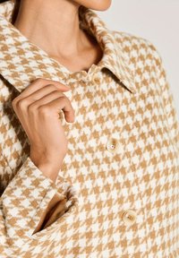 Vrouw in een beige en witte houndstoothgepatroneerde jas, die met haar linkerhand de kraag bij haar hals vasthoudt, met knopen zichtbaar aan de voorkant.