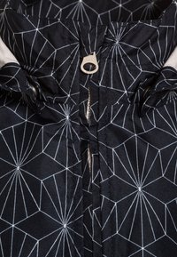 Giacca in tessuto nero con un motivo geometrico bianco, dotata di una chiusura a zip sul collo e di un cursore YKK, che mette in evidenza una texture e una struttura lisce.