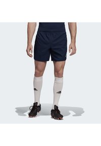 Marineblaue Sportshorts mit einem elastischen Bund, kombiniert mit weißen, kniehohen Socken mit einem schwarzen Logo, und schwarzen Fußballschuhen mit orangefarbenen Akzenten.
